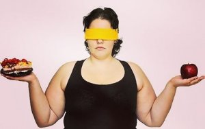 Mulher obesa com camiseta preta e olhos vendados com braços erguidos tem uma maça em uma mão e uma torta na outra.