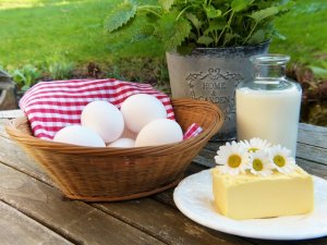 Mesa com cesto com ovos, prato com manteiras, flores margaridas e garrafa de vidro com leite.