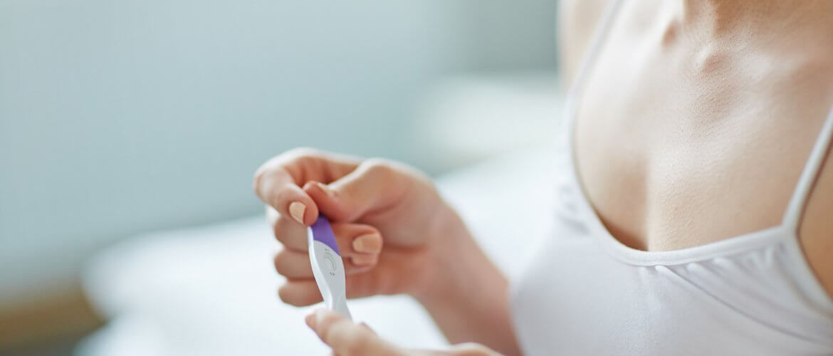 Mulher adepta do coito programado com camiseta de alças brancas segurando um teste de gravidez nas mãos.