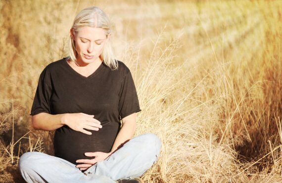 Mulher com gravidez tardia, sentada no campo, com mãos na barriga.